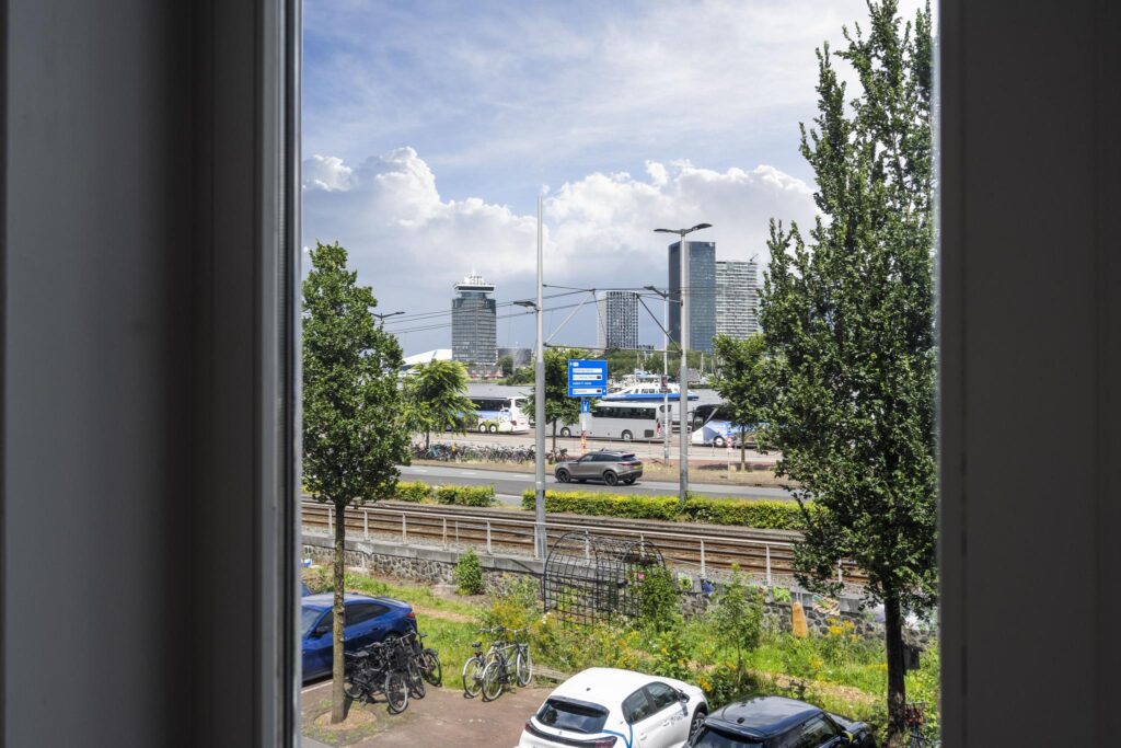 Uitzicht vanuit een raam op de De Ruijterkade in Amsterdam met geparkeerde auto's, treinspoor, bomen en moderne hoogbouw op de achtergrond.
